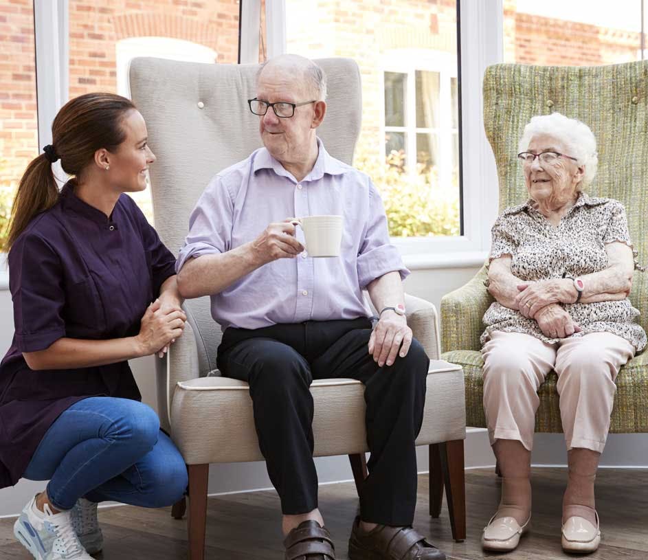 A caregiver kneeling and talking next to a smiling couple enjoying coffee